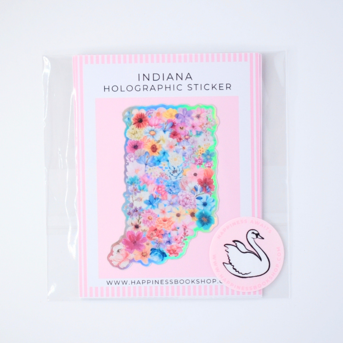 A packaged holographic sticker shaped like the state of Indiana, covered in a multicolor floral pattern, shown on a white background with a pink swan sticker also visible.