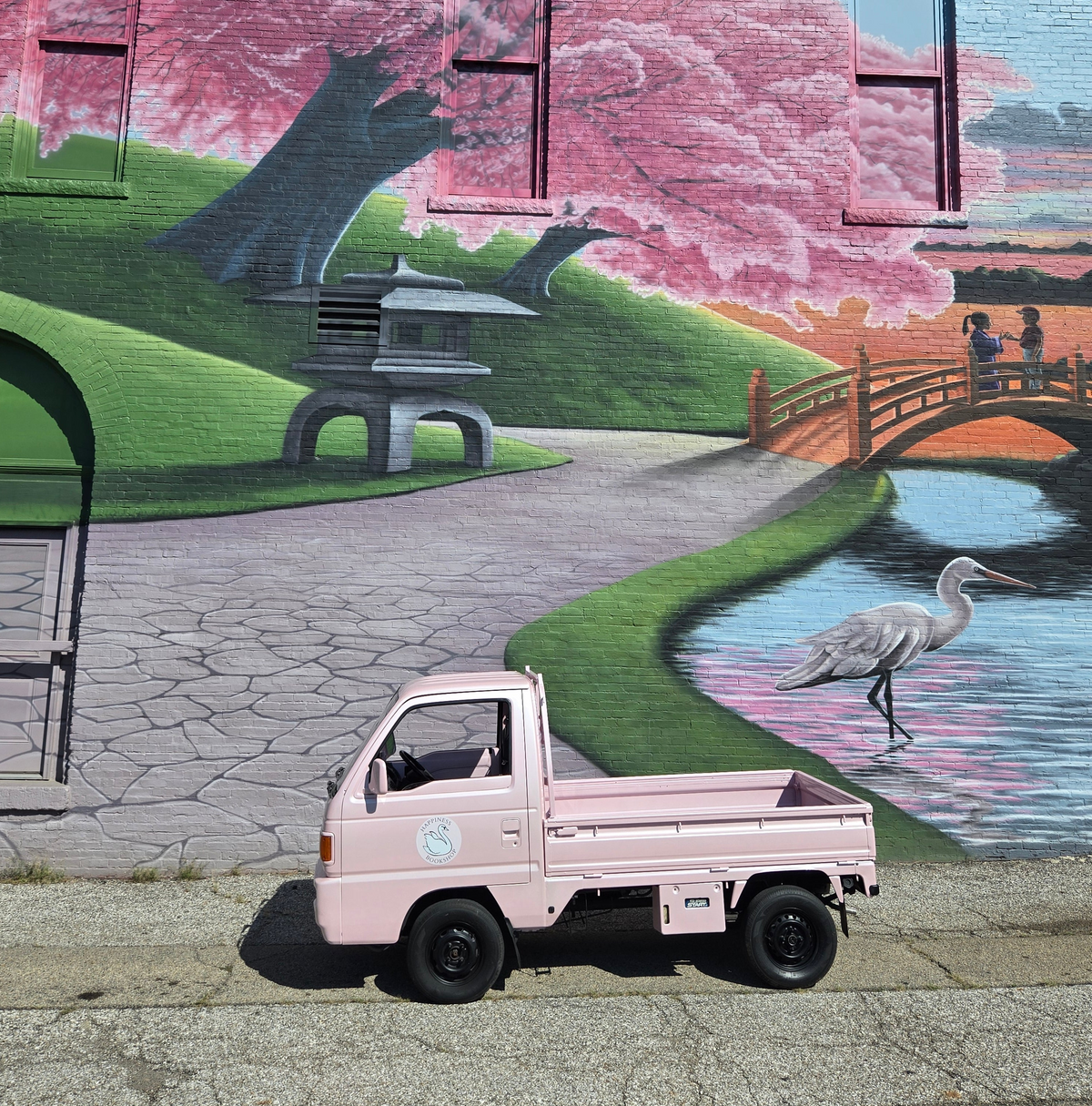 A pink miniature pickup truck parked on a street in front of a large, vibrant mural depicting a cherry blossom tree, a heron, a bridge, and a stone lantern.